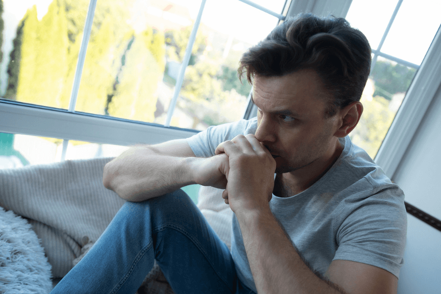 Man sitting by a large window with his hands clasped near his mouth, looking thoughtful and concerned, suggesting a moment of deep reflection or emotional struggle.