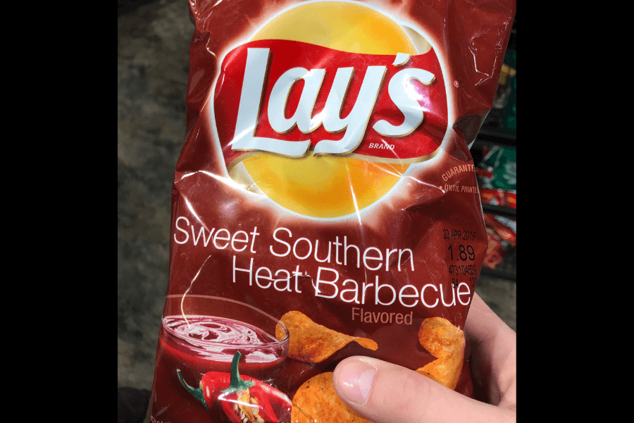 A bag of Lay's chips in the "Sweet Southern Heat Barbecue" flavor, featuring a rich brown design. The packaging displays an image of barbecue sauce with a chili pepper and seasoned chips to emphasize the sweet and spicy flavor profile. The bag is being held in a store, with shelves visible in the background.