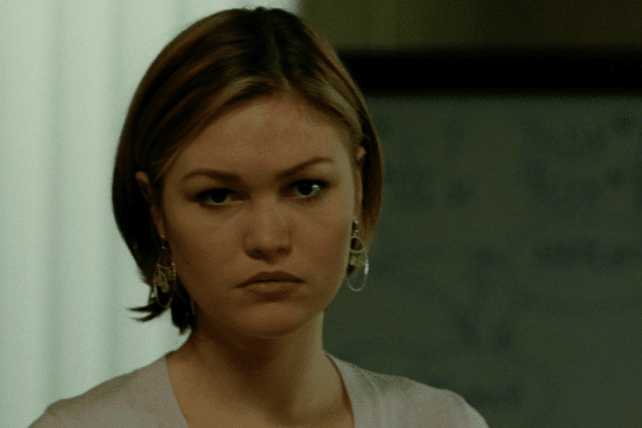 This image features a young woman with short, light brown hair, wearing earrings and a light-colored top. Her expression is serious or contemplative, and the background includes a blurred indoor setting with a whiteboard or similar object. The lighting and her demeanor suggest a moment of intensity or focus.
