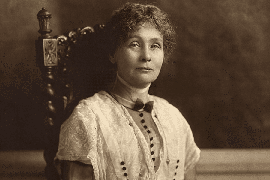 A sepia-toned portrait of a woman seated in an ornately carved wooden chair, wearing a high-collared lace blouse with button details. Her expression is calm and resolute, with short, wavy hair framing her face. The softly lit background emphasizes her poised and dignified presence.