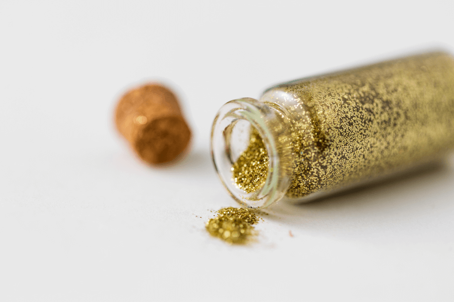 This image shows a small glass bottle tipped over on a white surface, spilling fine golden glitter. The cork lid is placed nearby, adding to the aesthetic of a delicate and creative craft setup. The focus on the glitter creates a sparkling, eye-catching effect.