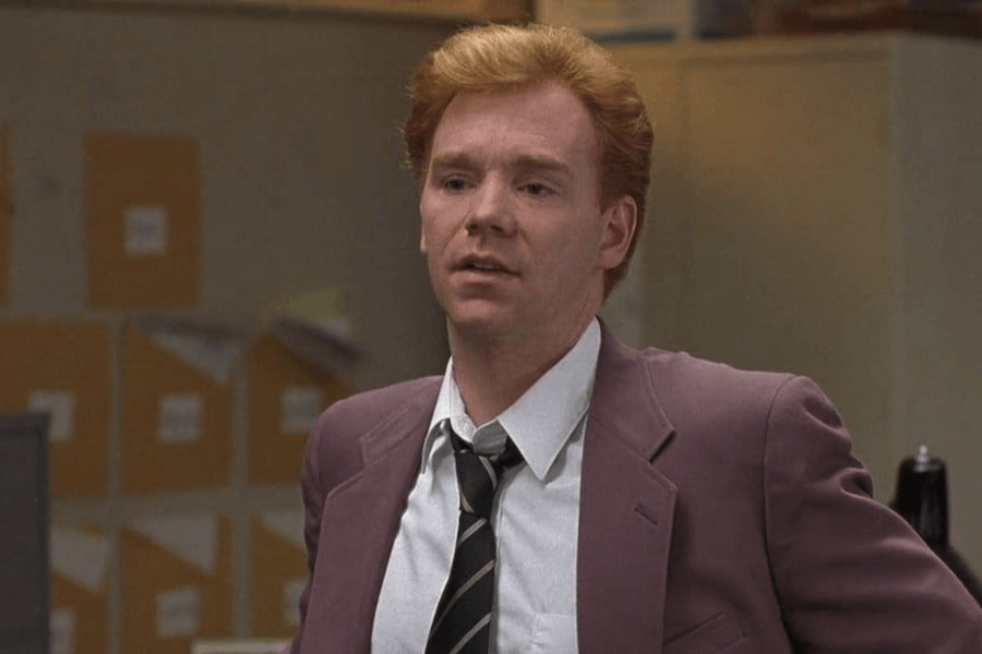 This image shows a man with reddish hair wearing a mauve-colored suit, a white shirt, and a striped tie. He appears to be in an office setting, with filing cabinets and boxes in the background. His expression is thoughtful or slightly weary, suggesting he may be engaged in a discussion or deep in thought.