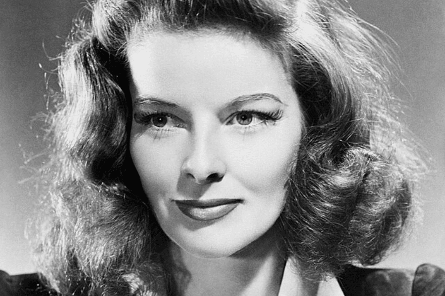 A classic black-and-white portrait of a woman with voluminous wavy hair and a poised expression. Her eyes are bright, and she has well-defined eyebrows, long lashes, and a slight smile. The dramatic lighting accentuates her facial features, giving the image a timeless Hollywood glamour.