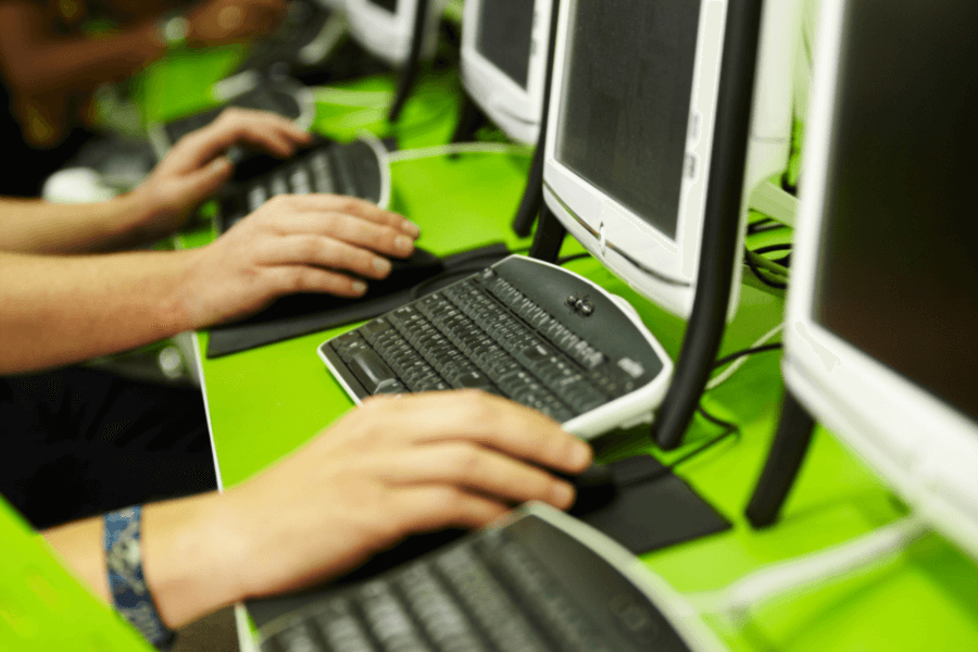 This image shows multiple hands typing on keyboards in a row of computers, set on a bright green desk. The setup suggests a shared workspace, computer lab, or internet café environment. The visible focus on screens and keyboards emphasizes collaborative or individual digital work.
