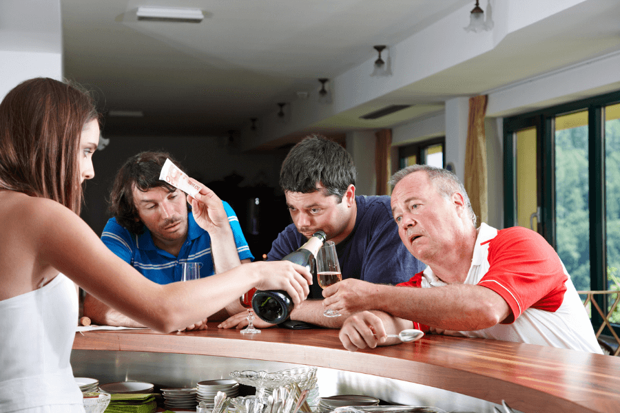 Three men at a bar appear intoxicated, with one holding a bottle, another offering money, and the third leaning heavily on the counter. A female bartender interacts with them while maintaining a professional demeanor in a brightly lit, casual setting.