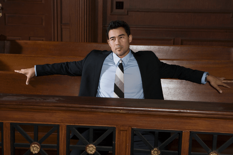 Man in a suit sitting confidently in a courtroom pew with arms stretched across the backrest, giving a relaxed yet assertive posture in a formal legal setting.