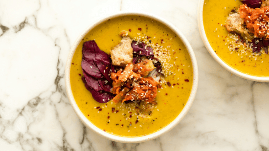 Two bowls of soup on a marble table with spoons and herbs