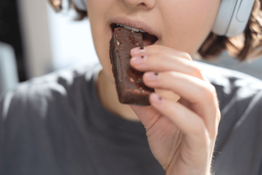 A close-up of a person taking a bite of a chocolate-covered snack bar while wearing headphones. Their nails are slightly chipped, and they are dressed in a casual gray t-shirt, with a blurred background suggesting a relaxed environment.