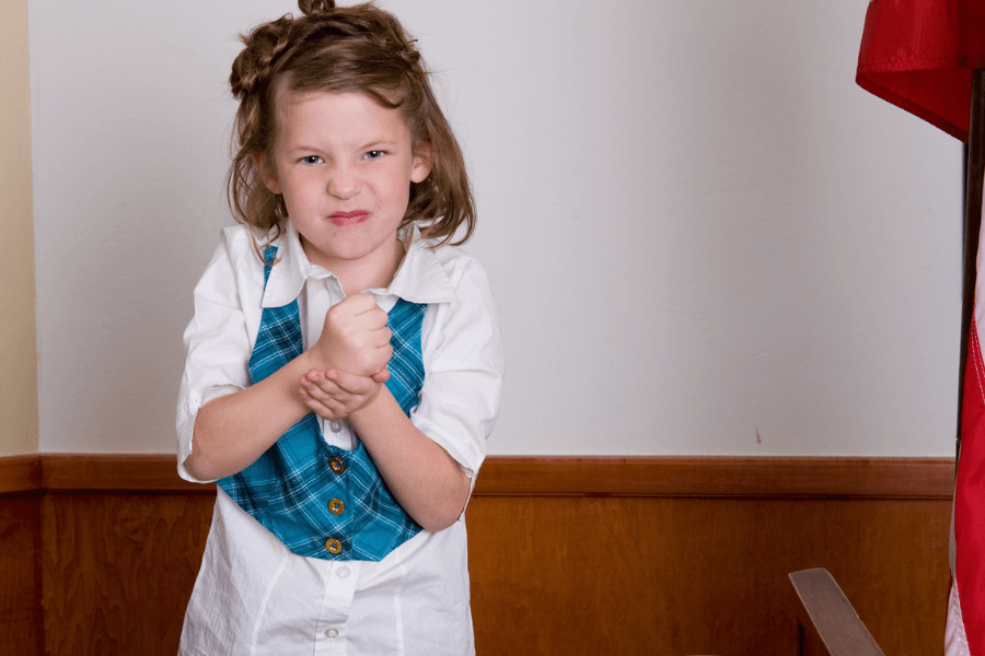 Young girl with a determined or mischievous expression, clenching her fists and wearing a teal plaid vest over a white shirt, standing in what appears to be a formal setting with a wooden panel and a red flag visible.