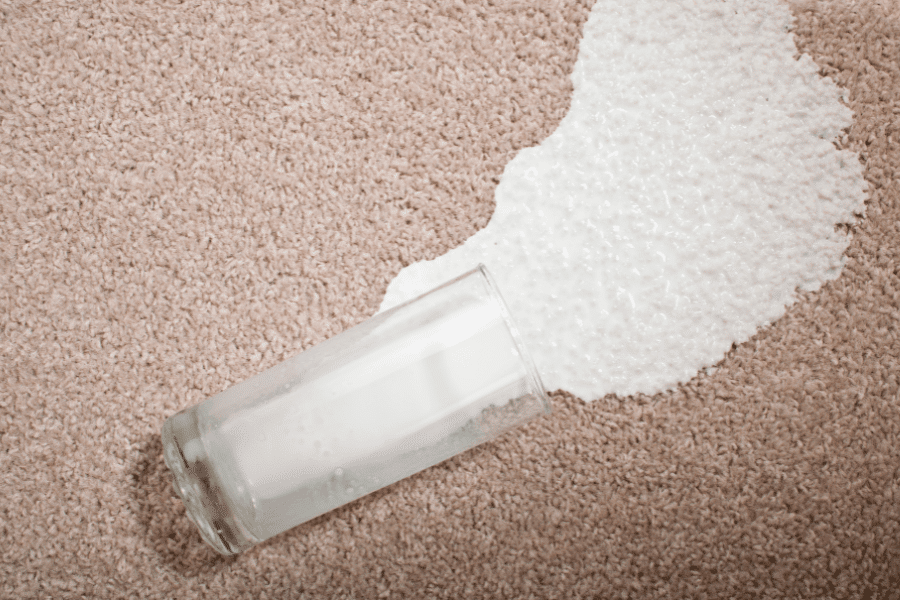 This image shows a glass of milk tipped over on a beige carpet, with milk spilled across the surface. The texture of the carpet and the contrast of the white milk emphasize the accidental spill. The scene captures a relatable moment of mishap.
