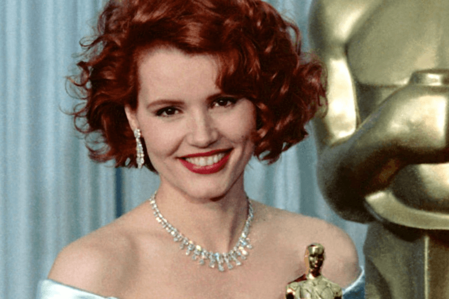 This image features a woman with short, curly red hair, dressed elegantly in a light-colored off-the-shoulder gown, accessorized with a sparkling necklace and earrings. She is smiling warmly while holding an Oscar trophy, with a large golden Oscar statue partially visible in the background, set against a blue curtain. The scene exudes glamour and celebration.