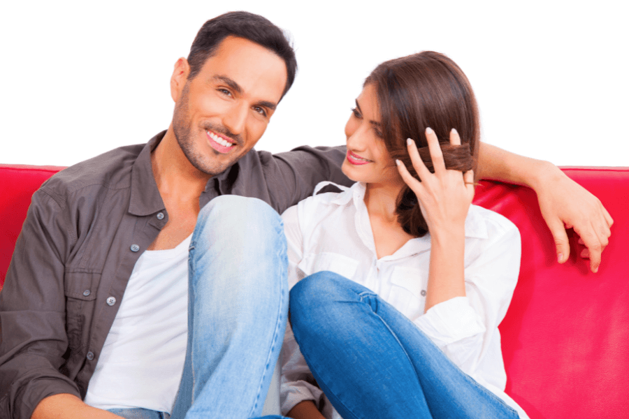 This image shows a smiling couple sitting together on a red couch. The man is casually dressed in a gray shirt and white undershirt, while the woman wears a white blouse and jeans, playfully touching her hair as she looks at him. The bright background highlights their cheerful interaction.