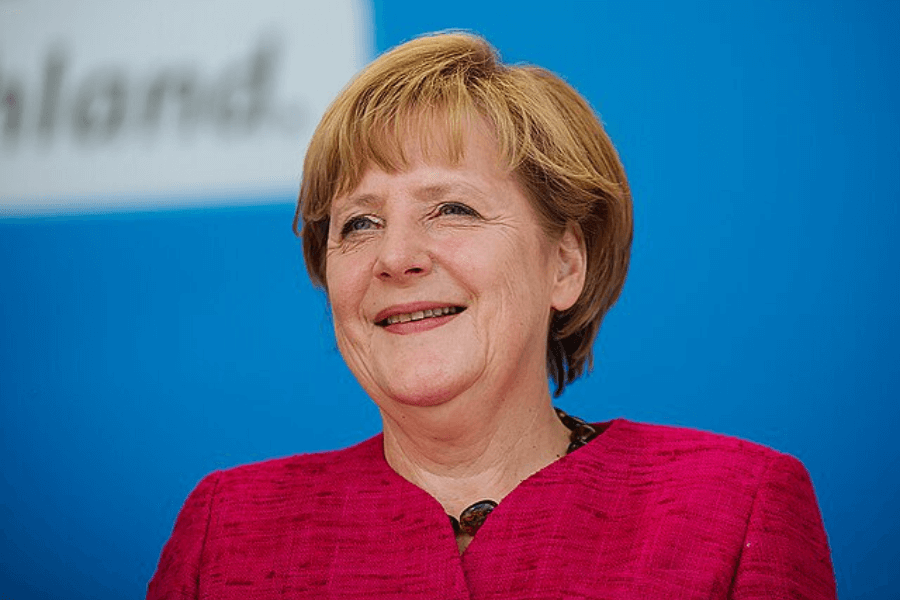 A woman with short blonde hair and a warm smile is wearing a bright pink blazer while standing against a blue background. She appears confident and approachable, with a relaxed posture. The background features partially visible text, suggesting a formal or public event setting.