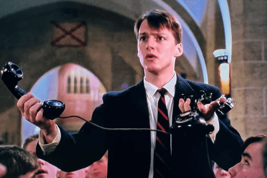 This image features a young man in a formal suit with a striped tie, standing in what appears to be a chapel or large hall. He is holding a disassembled black rotary phone with its parts spread in both hands, and his expression is one of surprise or determination. The background shows arched windows and a crowd seated around him, suggesting a dramatic or pivotal moment.