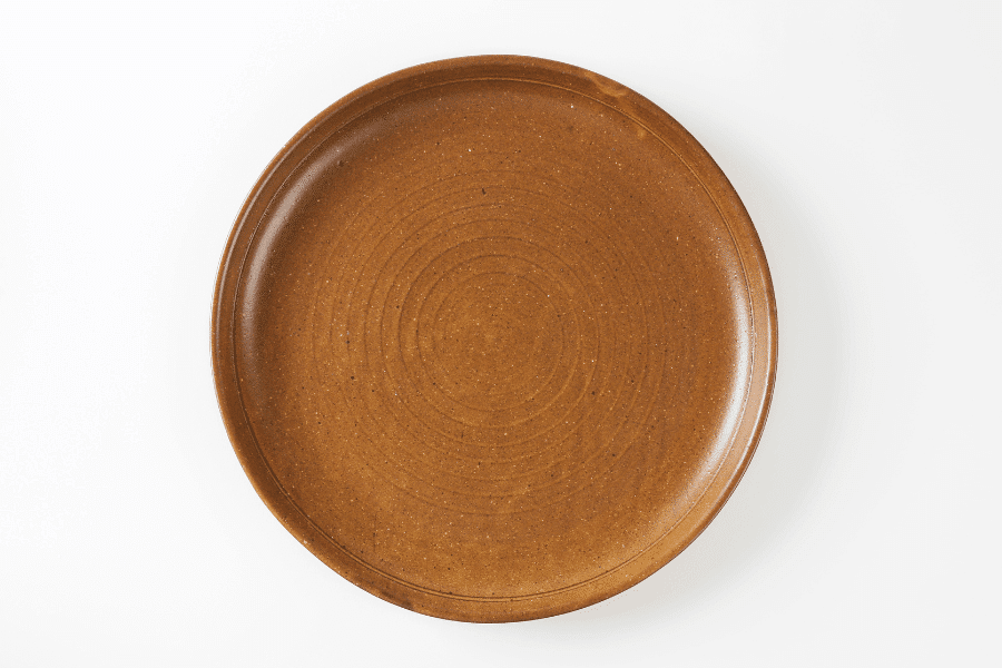 This image displays a round, brown ceramic plate with a minimalist design and concentric circular patterns. The plate has a matte finish and is set against a clean, white background, emphasizing its earthy tone and texture.