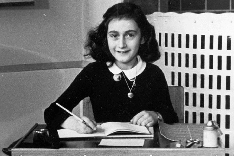 A black-and-white photograph of a young girl with dark, wavy hair sitting at a school desk, smiling as she writes in a notebook with a pencil. She wears a dark sweater with a white collared shirt underneath and a necklace with two small pendants. The setting appears to be a classroom or study area, with a heater visible in the background.