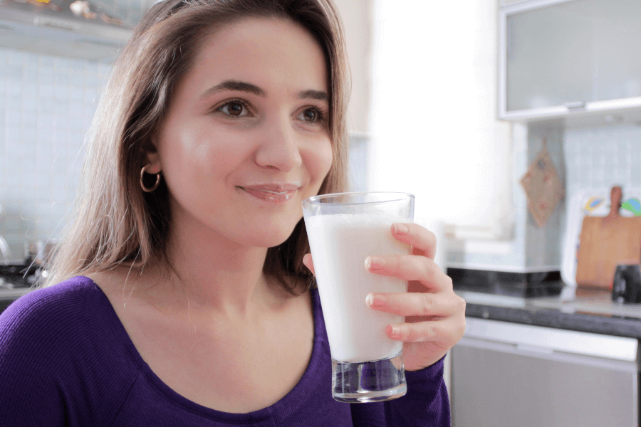 A smiling woman with long brown hair and gold hoop earrings holds a glass of milk in a bright kitchen. She wears a purple top and appears content while looking off to the side.