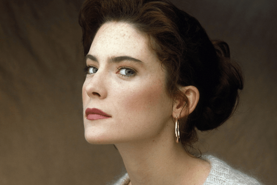 This image shows a woman with a poised and confident expression, looking directly at the camera from a slightly turned angle. She has auburn hair neatly tied back, accentuating her striking blue eyes and wearing subtle makeup. A pair of gold hoop earrings completes her elegant and classic appearance against a neutral backdrop.