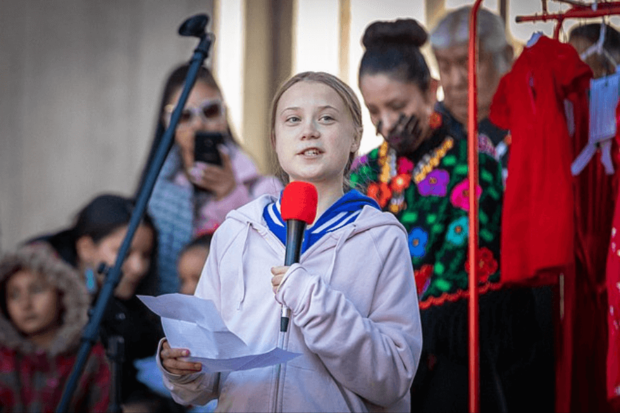 A young girl with light brown hair pulled back speaks into a microphone with a red cover while holding a piece of paper. She is wearing a light pink hoodie with a blue shirt underneath, addressing a crowd at an outdoor event. Behind her, several people in colorful and traditional clothing are present, some watching attentively while others are engaged in different activities.