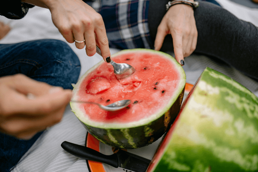 A group of people scooping out fresh watermelon with spoons, eating directly from the halved fruit. The vibrant red flesh contrasts with the green rind, and a knife rests on an orange plate nearby.