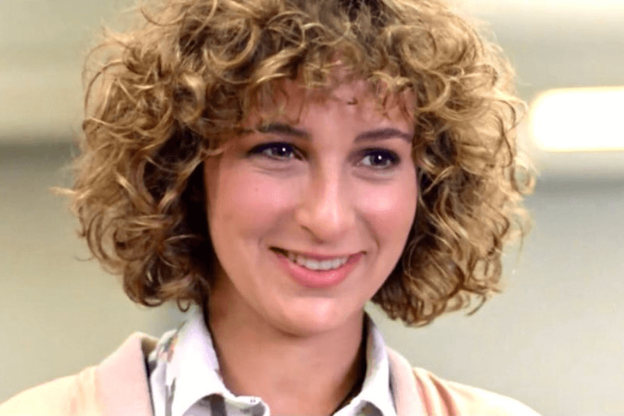 This image shows a smiling woman with curly, shoulder-length hair and a casual, light-toned outfit. Her warm expression and soft lighting create an inviting and cheerful mood, complemented by a plain indoor background.