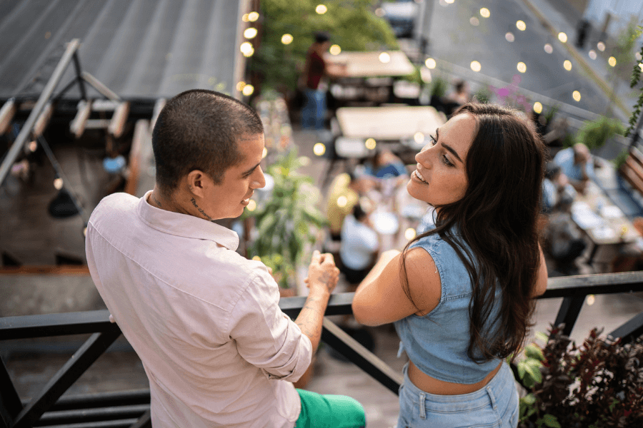 Young couple enjoying a relaxed conversation on a rooftop terrace, surrounded by string lights and outdoor dining tables, with a lively city ambiance in the background.