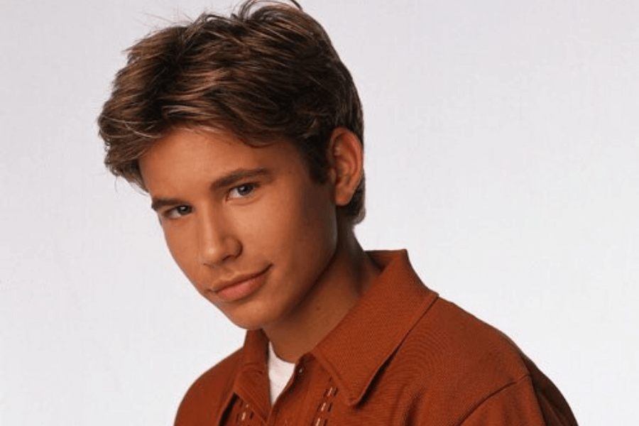 This image features a young man with styled light-brown hair and a confident expression, wearing an orange collared shirt. The background is a simple white, emphasizing a clean and professional portrait-style composition.