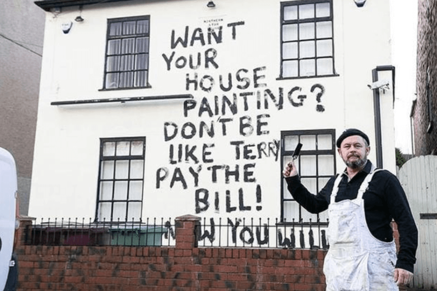 This image features a man wearing painter's overalls standing in front of a house. The wall of the house has large, bold, and unevenly painted text that reads: "Want your house painting? Don't be like Terry. Pay the bill!" The man is holding a paintbrush and appears to be pointing to the message, which is likely a humorous act of revenge for non-payment.