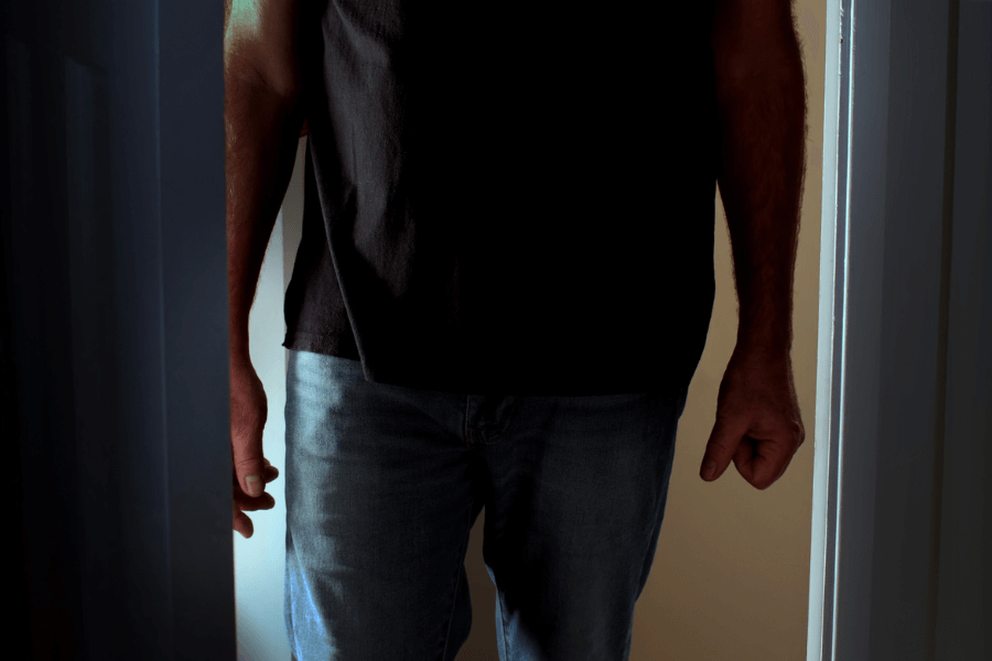 Partial view of a man standing in a doorway with clenched fists, wearing a black shirt and jeans, illuminated by dim lighting that creates a tense, possibly confrontational atmosphere.