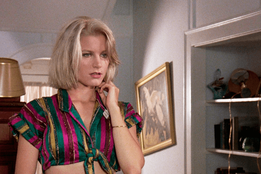 This image shows a woman with a blonde bob haircut, wearing a colorful striped crop top tied at the waist. She appears thoughtful or pensive, with her hand near her chin, standing in a room with light-colored walls, framed artwork, and shelves in the background.