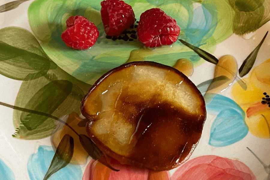 This image shows a decorative plate featuring a caramelized slice of fruit, likely an apple, drizzled with syrup and accompanied by three fresh raspberries. The vibrant plate design with floral patterns adds a colorful and artistic backdrop to the dessert-like presentation.