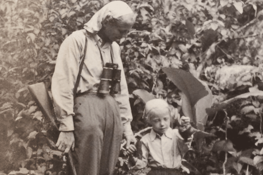 Black-and-white photo of an older woman and a young blonde child in a dense jungle. The woman wears a headscarf, has binoculars around her neck, and carries a rifle over her shoulder. The child looks curiously at a large leaf they are holding.