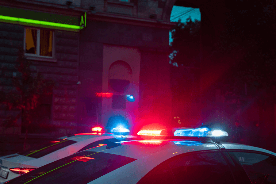 Two police cars with flashing red and blue lights parked on a dimly lit urban street at night, with a stone building and neon signage visible in the background.