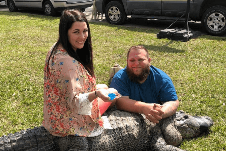 This image shows a woman and a man sitting on or near a restrained alligator outdoors on a grassy area. The woman is holding small blue objects in her hand, smiling at the camera, while the man sits beside her with a relaxed expression. Cars are visible parked in the background.