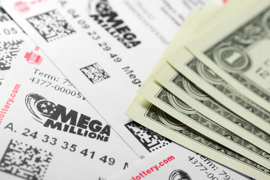 A close-up of multiple Mega Millions lottery tickets spread out on a surface, with a stack of U.S. dollar bills partially covering them. The tickets display the Mega Millions logo, QR codes, and sets of numbers. The image conveys themes of gambling, luck, and potential winnings.