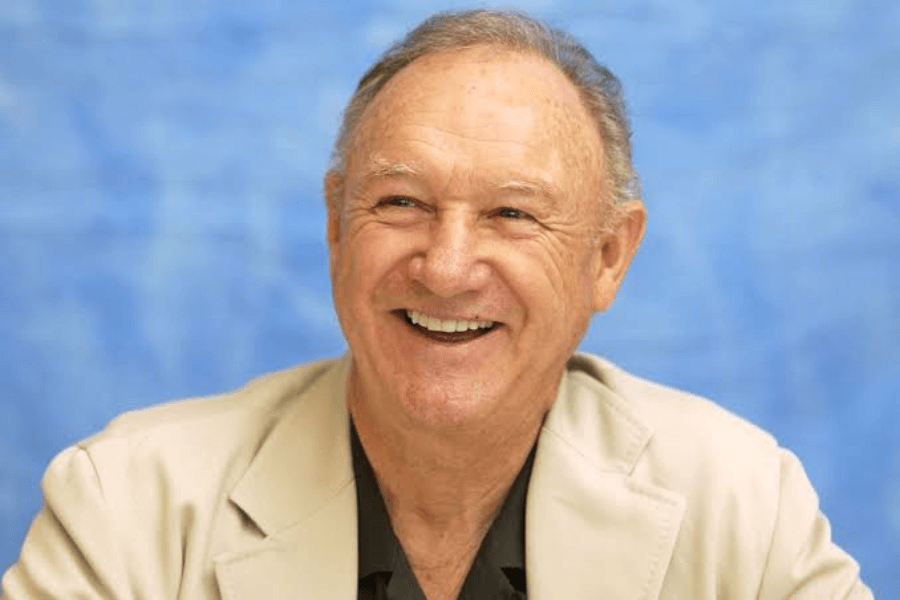 This image features an older man with a warm smile, wearing a beige blazer over a dark shirt. The background is a vibrant blue, creating a friendly and professional portrait-style composition.