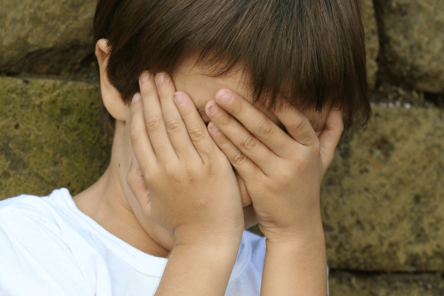This image shows a young child with their hands covering their face, possibly in shyness, embarrassment, or playfulness. The background features a stone wall, and the child is wearing a plain white shirt.