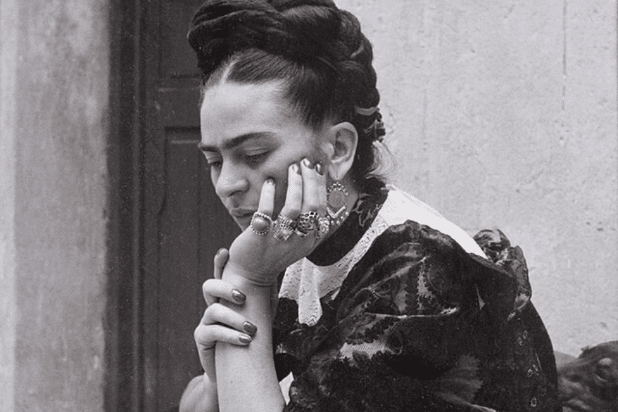 A black-and-white photograph of a woman with braided hair, resting her face on her hand in a contemplative pose. She wears ornate rings and earrings, along with a patterned blouse featuring lace details. Her gaze is directed downward, conveying a pensive mood against a simple background.
