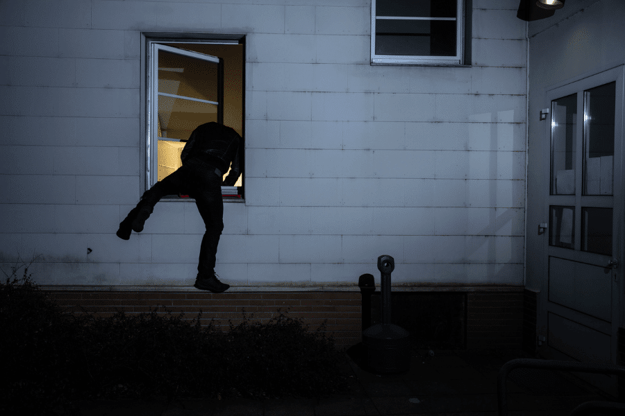 Person dressed in dark clothing climbing through a lit window into a building at night, suggesting a burglary or unauthorized entry.