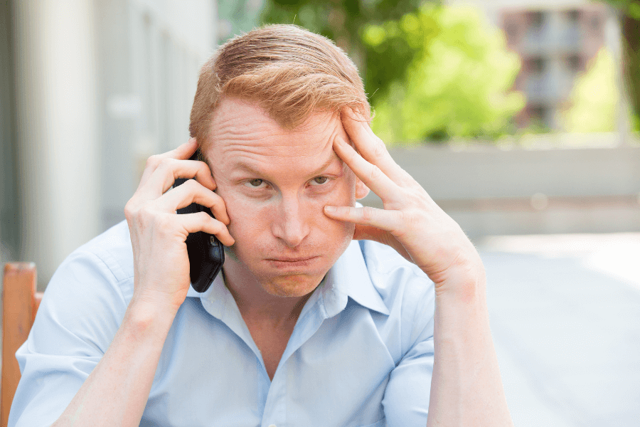 This image shows a man with a frustrated or exasperated expression, holding a phone to his ear with one hand while resting his other hand on his forehead. He is wearing a light blue collared shirt and is outdoors, with a blurred background of trees and buildings.
