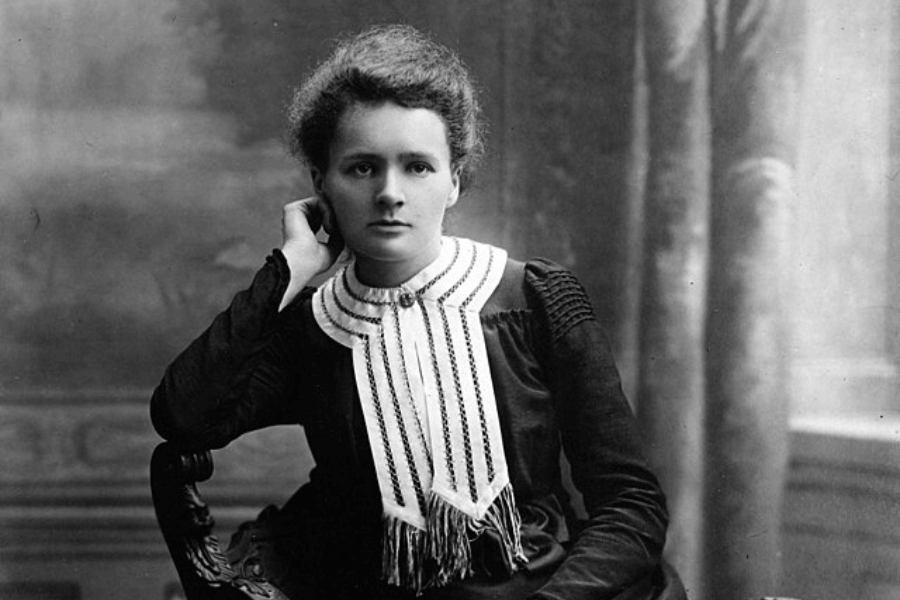 A black-and-white portrait of a woman seated in an elegant pose, resting her head on one hand while gazing directly at the camera. She wears a dark dress with a white, pleated collar adorned with delicate detailing. The background features a softly blurred, classical-style interior, adding to the formal and intellectual tone of the image.