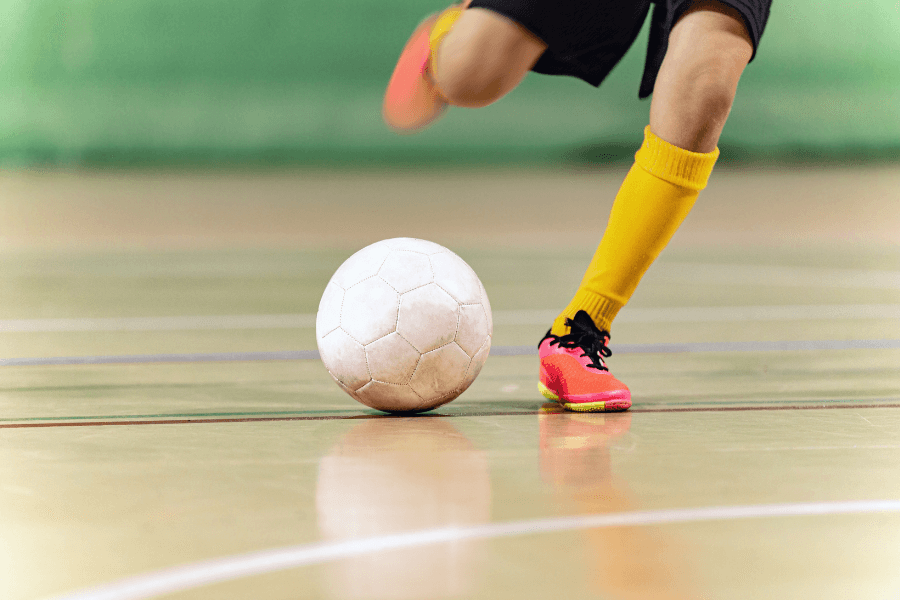 This image captures an indoor soccer scene where a player wearing yellow socks and bright pink cleats is in motion with a white soccer ball on a polished court. The blurred green background emphasizes the action and focus on the ball and player's legs.