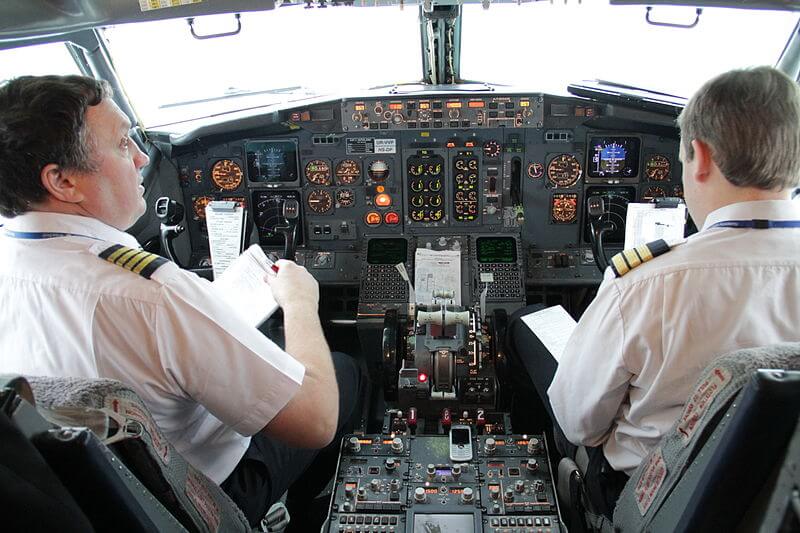 Two pilots in white uniforms with epaulets sit in the cockpit, reviewing checklists before flight. The control panel in front of them is filled with illuminated gauges, switches, and screens, creating a complex and focused work environment.