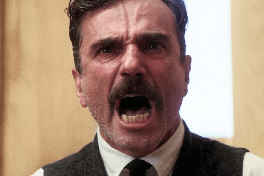 This image captures a man mid-shout, with an intense and emotional expression. He has a thick mustache, is wearing a suit with a tie, and appears to be in a brightly lit indoor setting with a wooden backdrop. The scene conveys anger or frustration with dramatic intensity.