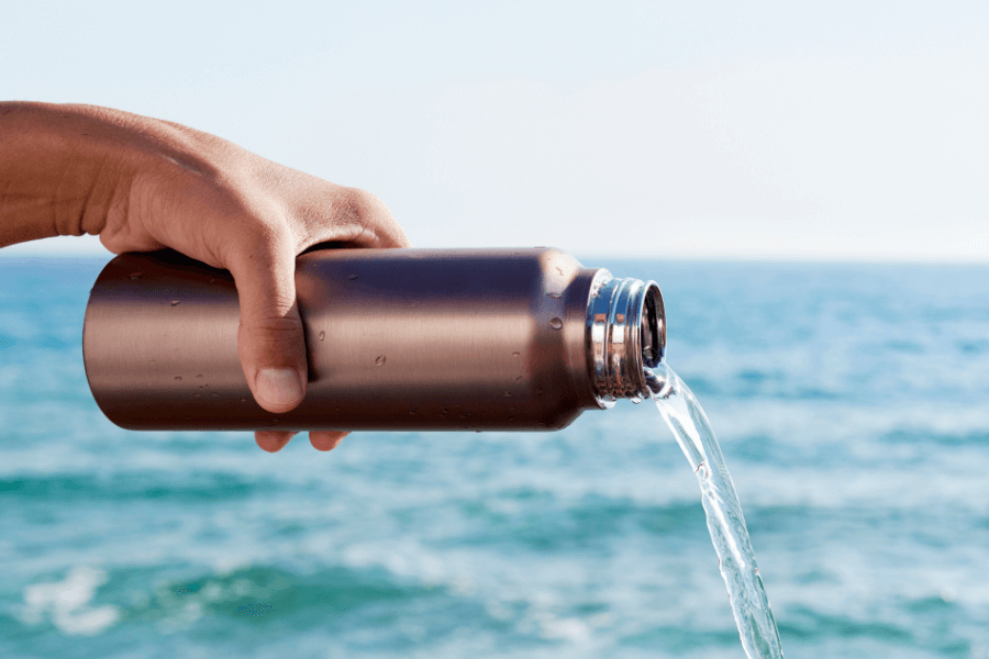 This image depicts a hand holding a metallic water bottle, pouring water out against the backdrop of a bright blue ocean and sky. The action and the serene setting evoke a sense of refreshment and connection with nature.