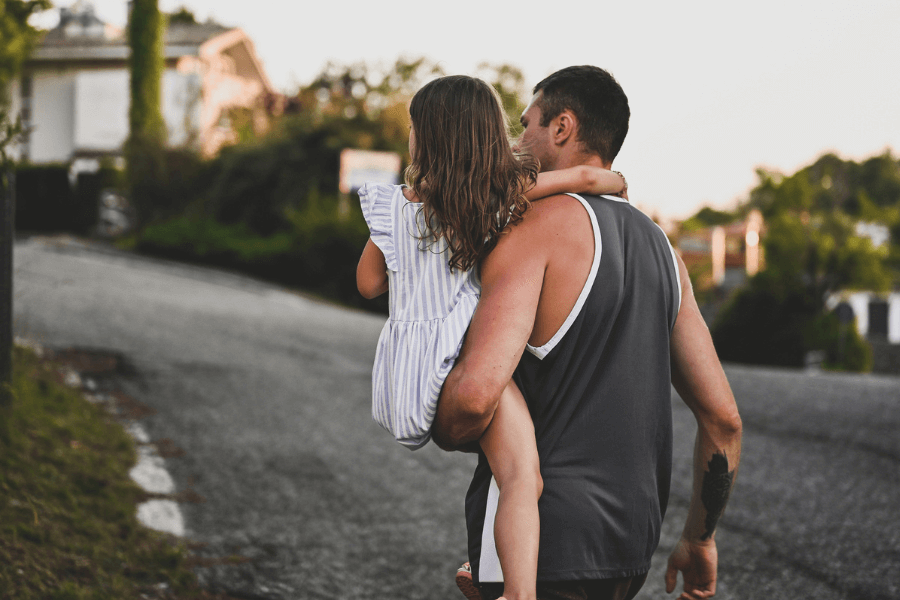 Man carrying a young girl in a striped dress as they walk up a quiet residential street at sunset, creating a warm and protective father-daughter moment.