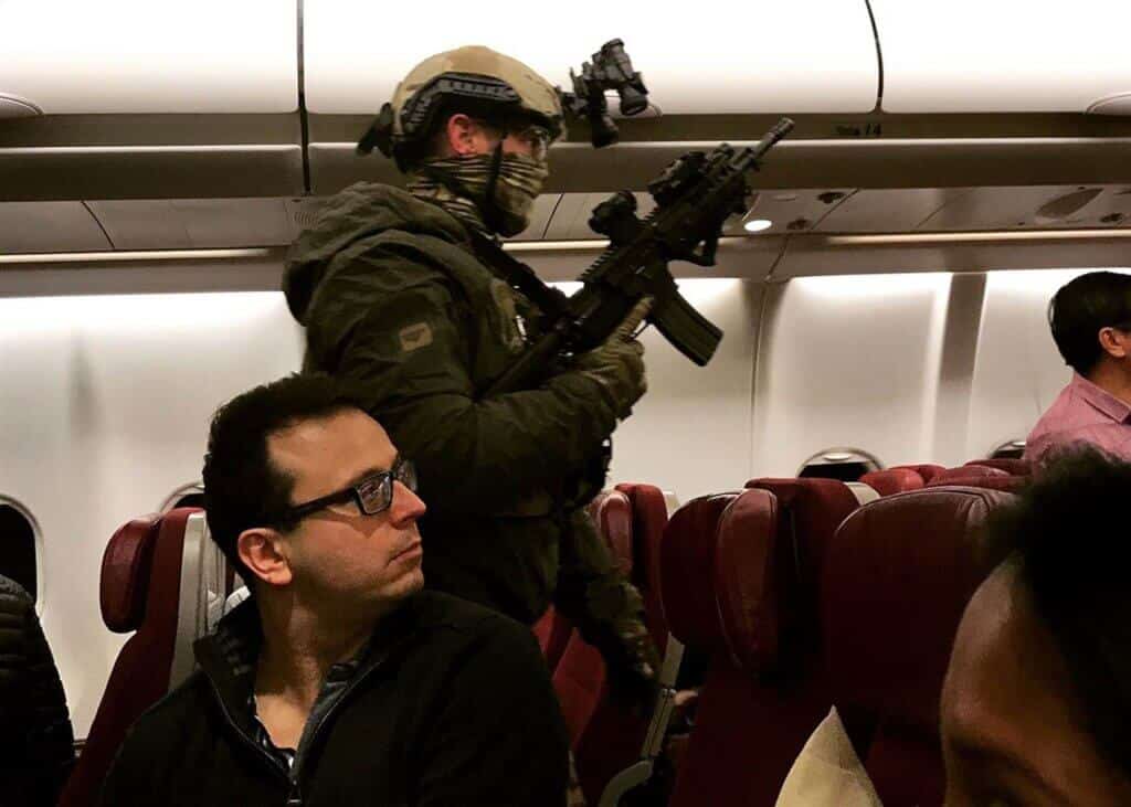 A heavily armed soldier in tactical gear, wearing a helmet and face covering, stands inside an airplane aisle holding a rifle. Passengers seated nearby glance cautiously, their expressions ranging from curiosity to concern in the dimly lit cabin.