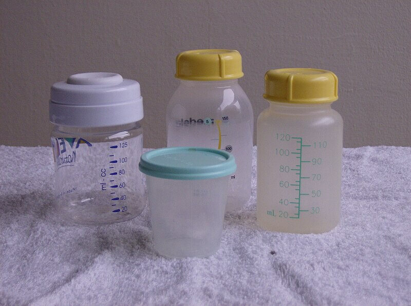 Four plastic baby bottles and storage containers of varying sizes and brands are placed on a white towel. Two bottles have yellow lids, one has a white lid, and the smallest container has a light blue lid, all marked with measurement indicators.