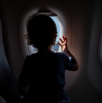 A young child, silhouetted against the light, gazes out of an airplane window with their hand reaching toward the glass. The soft glow of daylight outside contrasts with the dark interior, capturing a quiet moment of wonder and curiosity.