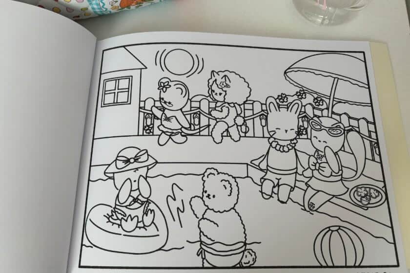 An open coloring book displays a black-and-white illustration of anthropomorphic animals enjoying a poolside scene. A bear in swim trunks splashes in the water while a penguin wearing a sunhat floats in an inflatable ring, surrounded by other cheerful characters lounging and socializing under the sun.